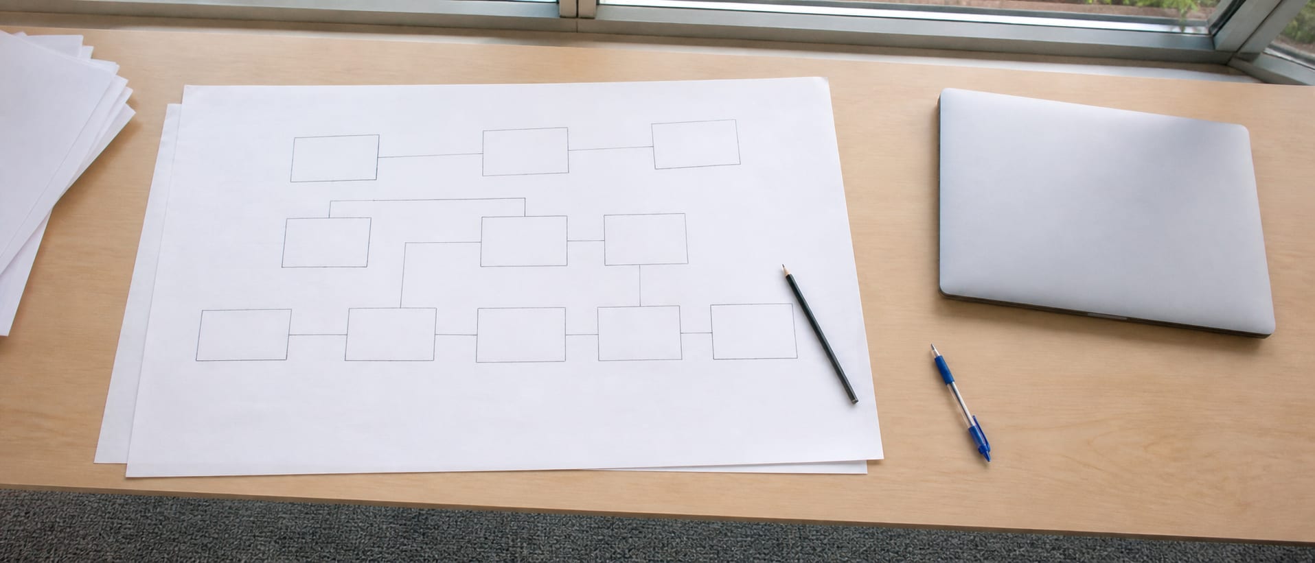 A wide desk carrying a hand-drawn workflow diagram on a long sheet of paper, boxes and arrows in pencil, with a laptop closed at one end