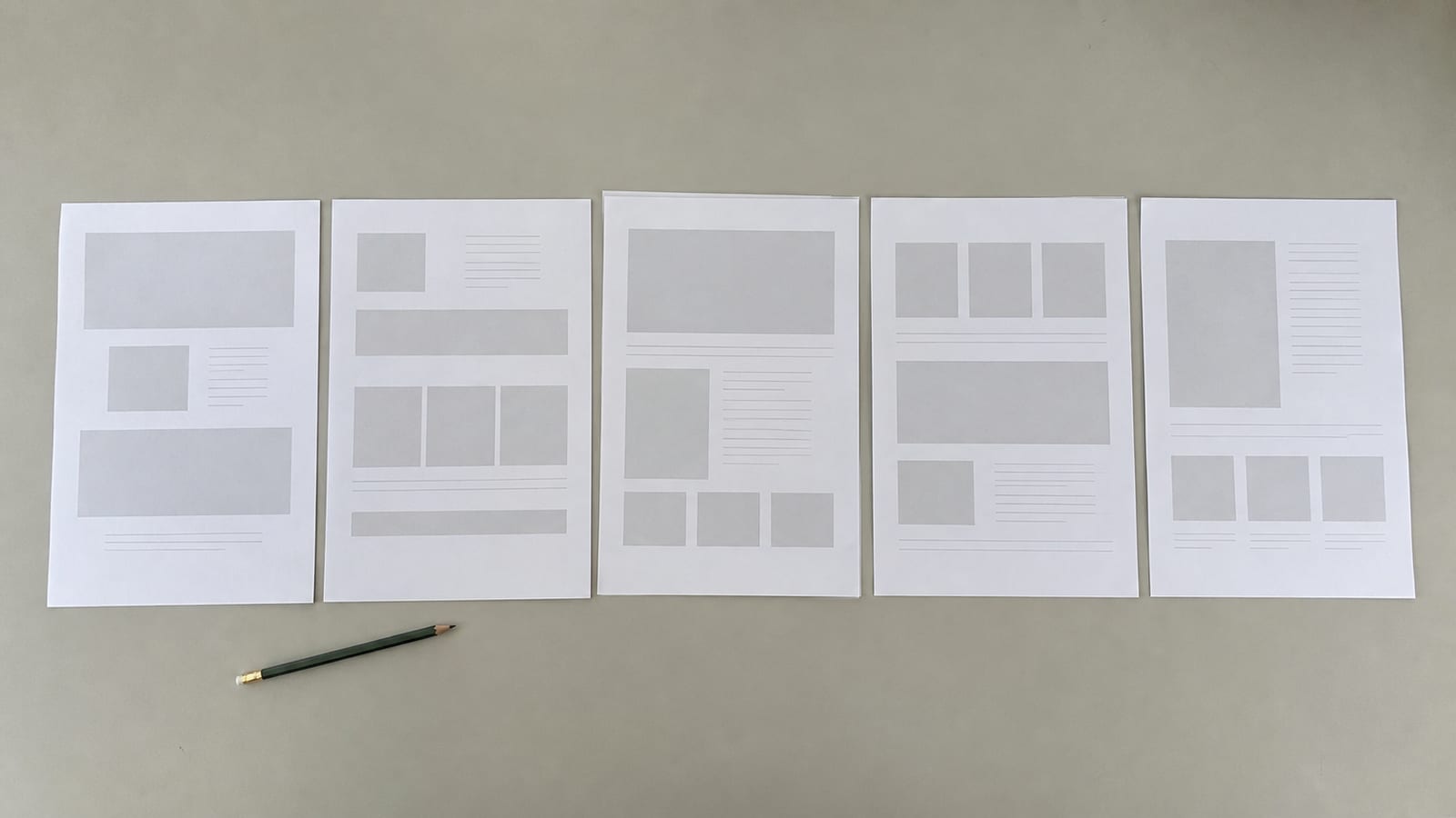 Five printed page proofs laid in a row on a plain surface, edges squared, each showing a different simple grey layout, seen from directly above