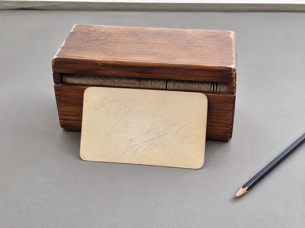 A single index card lifted clear and standing propped against the closed card box behind it, the card handwritten and softened at the corners from handling