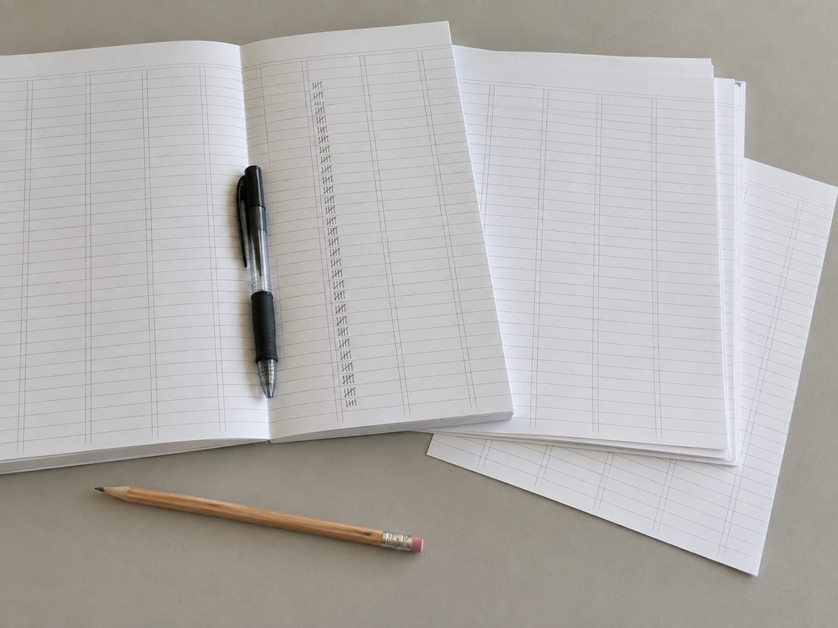 A printed ledger sheet on a desk ruled into narrow columns with a run of handwritten tally strokes down one column, a ballpoint resting in the gutter