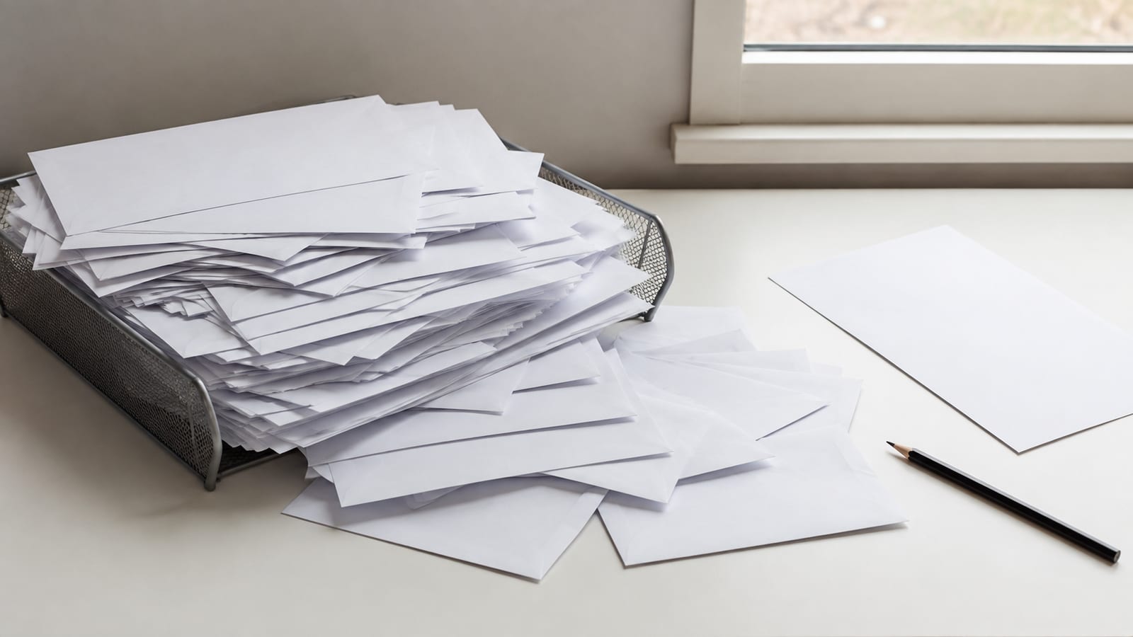 A mail tray on a desk overflowing with unopened envelopes, a further loose drift of envelopes spilling onto the desk beside it, all plain and unaddressed