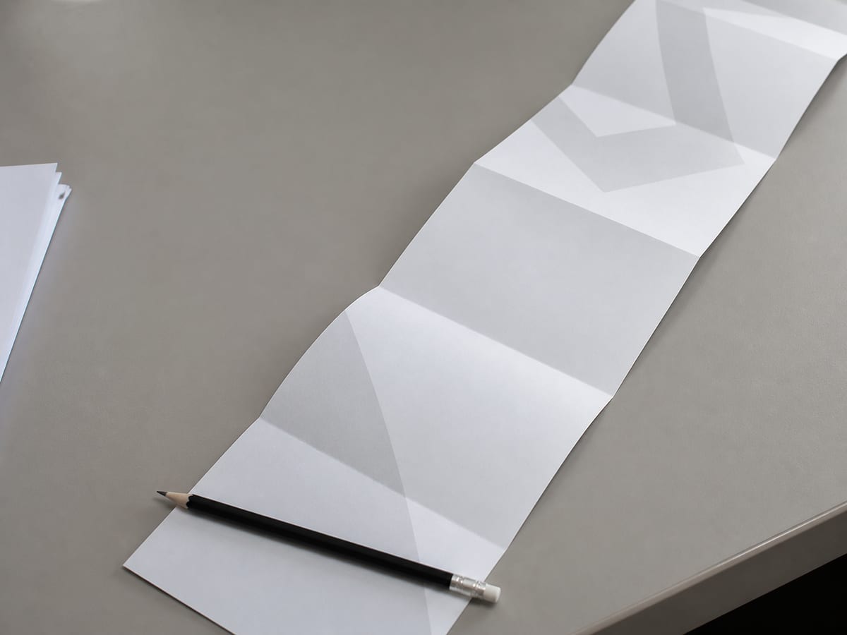 One printed leaflet unfolded to its full length so the far end runs off the edge of the desk, the near end weighted flat by a pencil