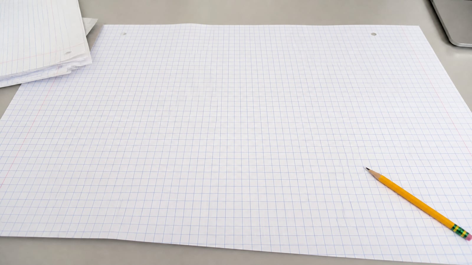 A large sheet of ruled analysis paper spread across a desk, far wider than the sheets around it, its grid extending past the frame on both sides, held flat at one corner by a pencil