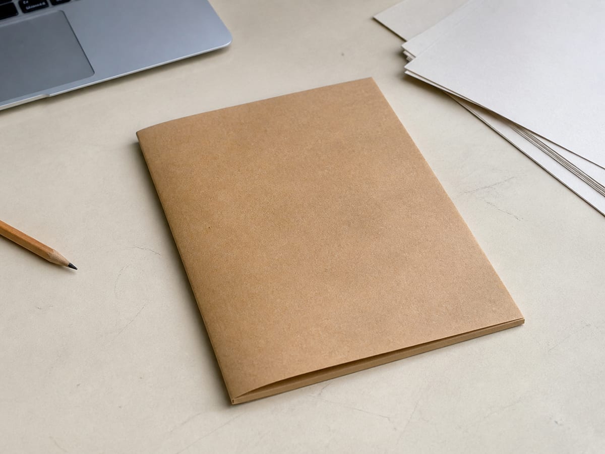 A buff paper wallet on a desk visibly aged and softened at the edges, closed, with a thin layer of dust across its upper face