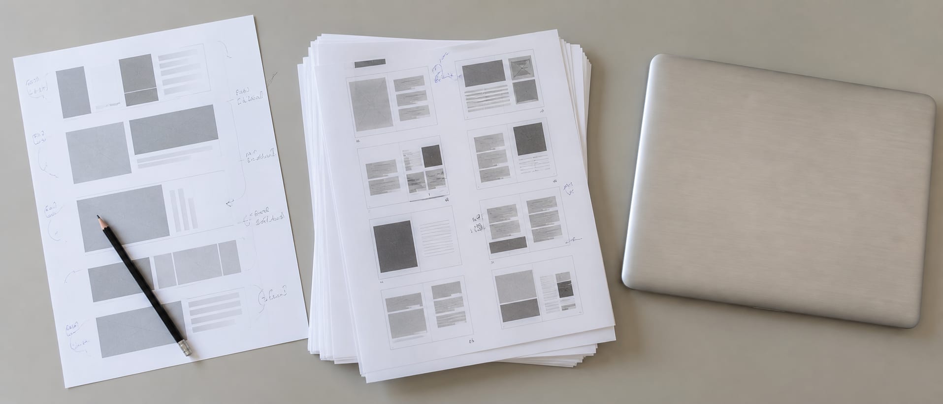 A wide desk surface seen straight down, carrying a printed page-structure diagram, a stack of printed page proofs squared up, and a closed laptop, arranged as a working layout rather than a styled flat lay